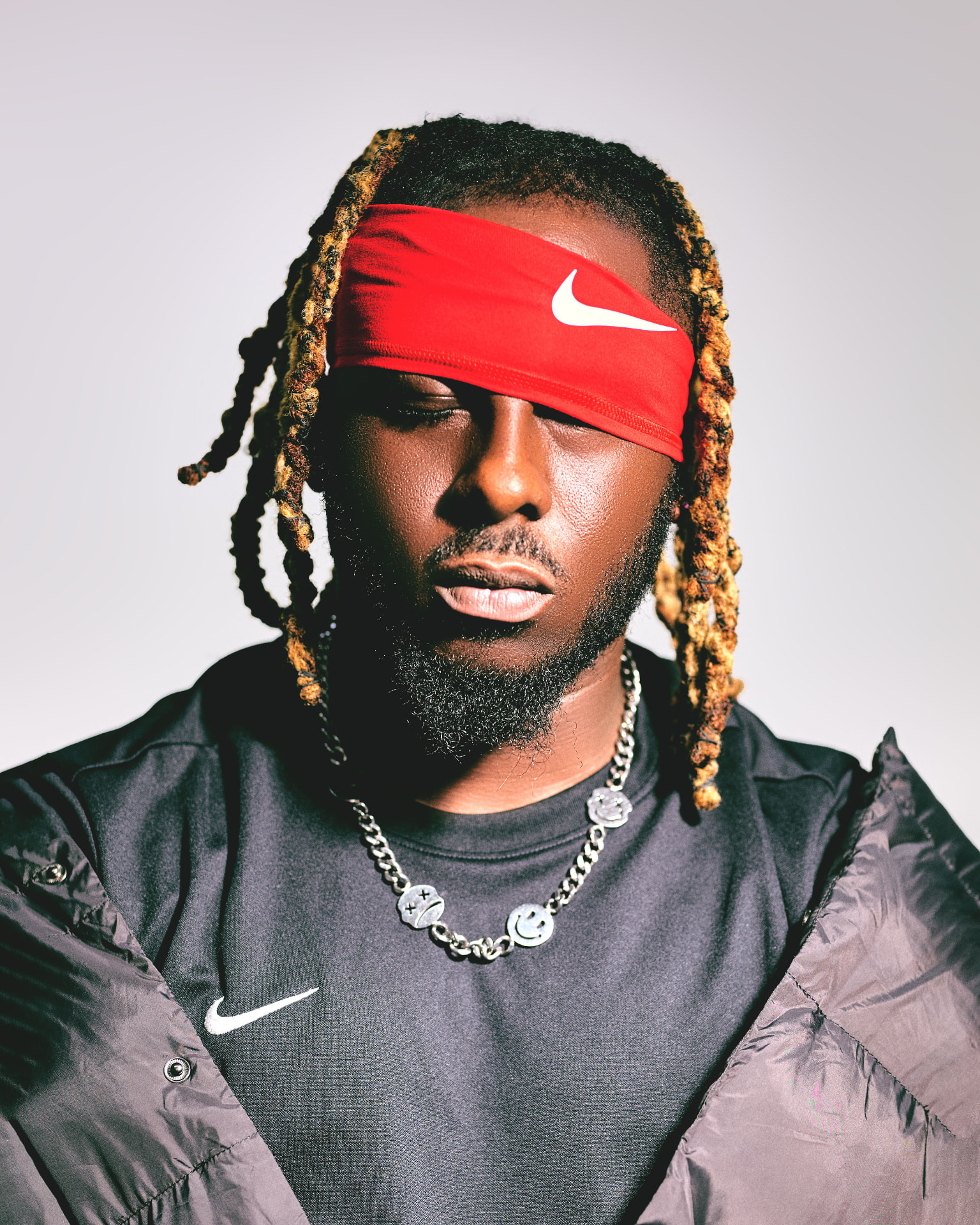 Max Meezy with a red headband pulled low over his eyes, gold-tipped dreadlocks and a silver chain, photographed against a pale studio wall